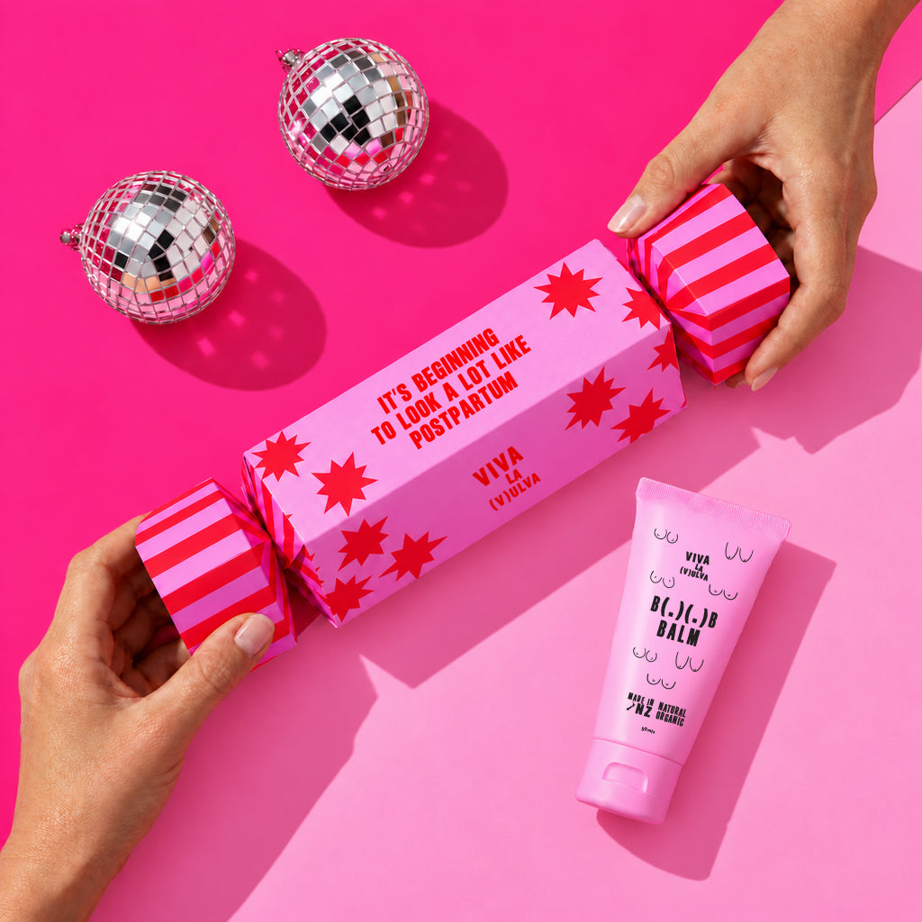 Hands opening a pink Viva La Vulva Christmas cracker gift box with a B( . )( . )B Balm nipple balm tube beside it