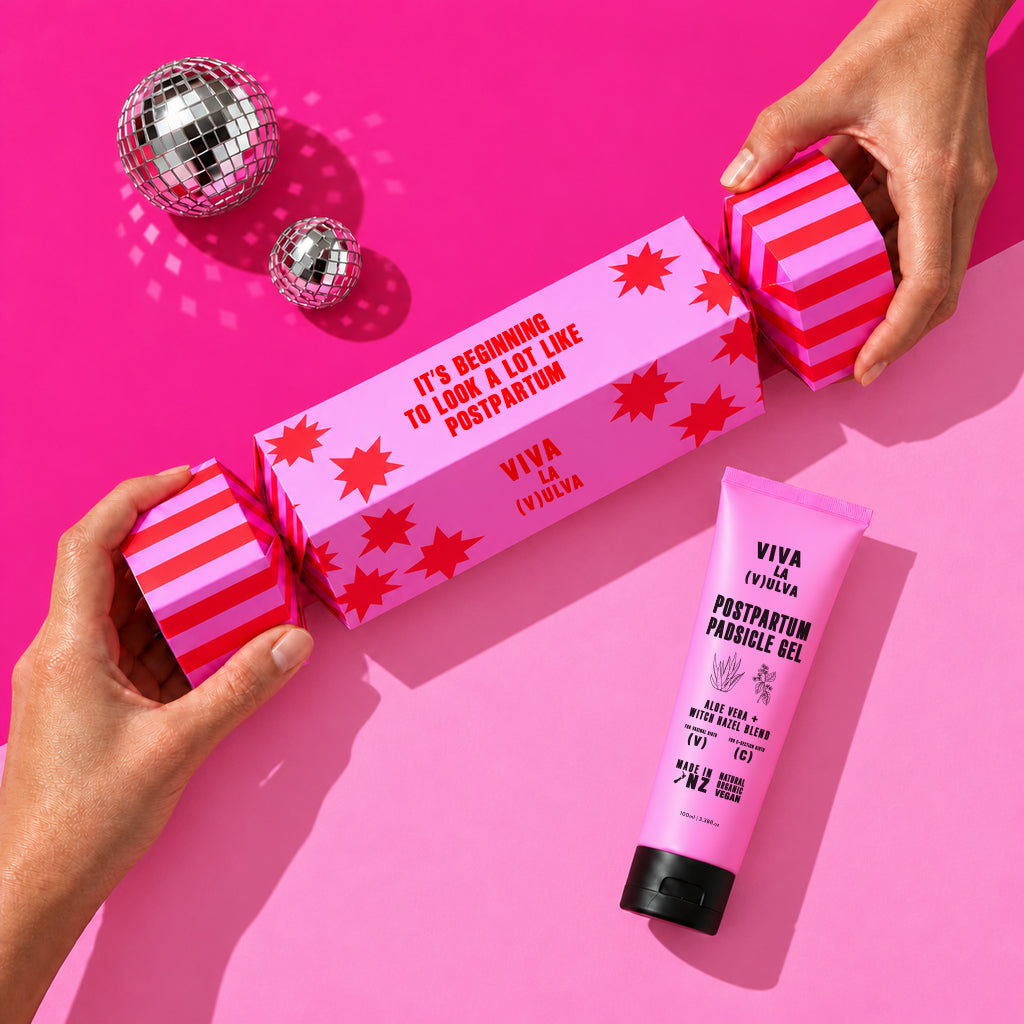Hands opening a pink Viva La Vulva Christmas cracker gift box with a Postpartum Padsicle Gel tube beside it